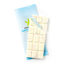 Unwrapped Kush Kitchen Birthday Cake THC Chocolate Bar. The white chocolate bar with colorful sprinkles is partially broken, revealing individual squares. The light blue packaging is visible underneath the chocolate bar