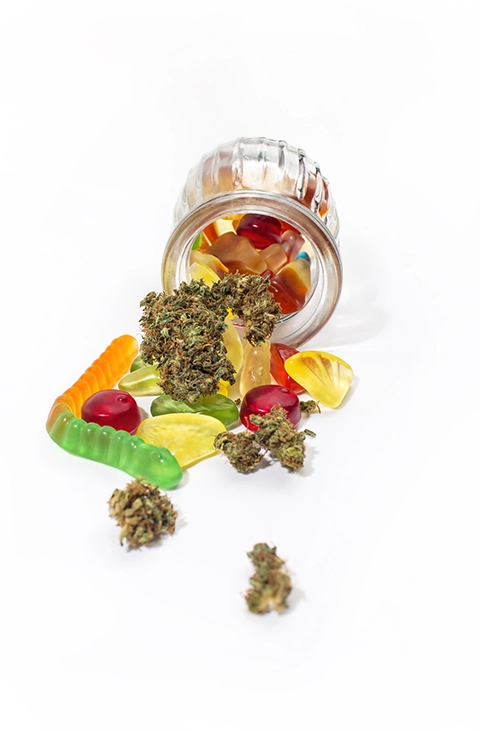 A glass jar spilling out buy edibles online gummy candies alongside cannabis buds, showcasing the delicious variety of cannabis edibles available for a convenient and enjoyable experience.