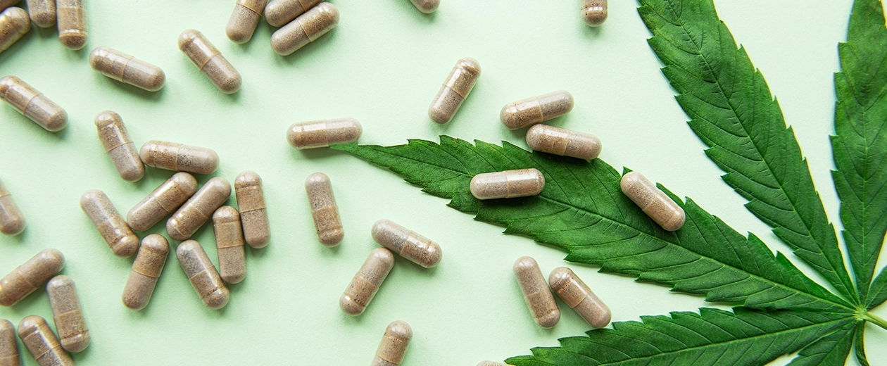 A collection of THC capsules scattered on a green surface alongside a fresh cannabis leaf, highlighting the natural and potent benefits of cannabis THC capsules for convenient consumption.