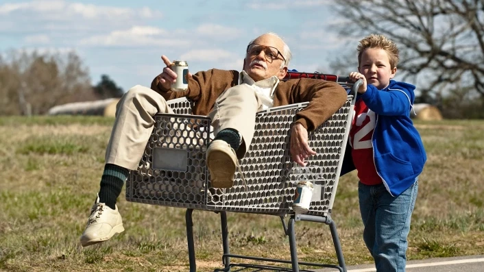 An old man rides in a shopping cart holding a beer while a young boy pushes him down a country road in Bad Grandpa