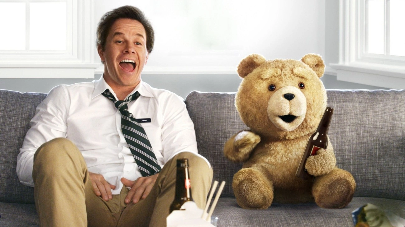 Mark Wahlberg laughs on a couch beside Ted the talking teddy bear, both holding beers